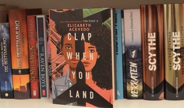 Buch "Clap When You Land" vor einem Regalbrett in einem gefüllten Bücherregal.